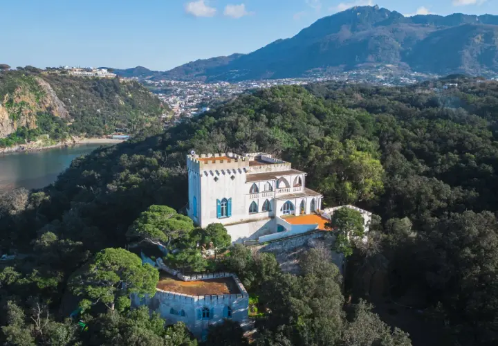 Villa La Colombaia a Ischia, un bianco maniero con torrette e finestre blu, immerso nel verde lussureggiante con vista sul mare e sulle montagne.