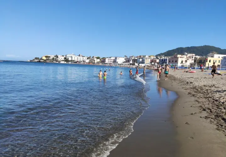 Persone si godono una giornata di sole sulla spiaggia di San Pietro, con il mare azzurro e case colorate sullo sfondo.