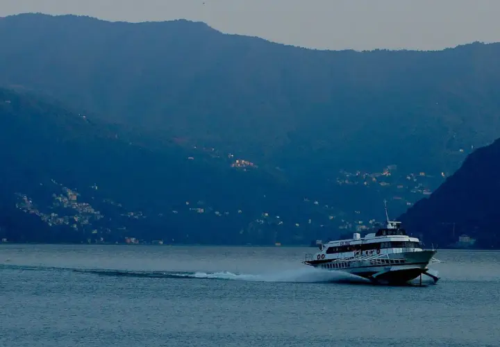 Aliscafo in navigazione su un lago calmo, con montagne boscose e case illuminate sullo sfondo.