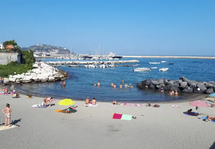 Spiaggia della Marina di Ischia: bagnanti si godono il sole e il mare, con barche ormeggiate nel porto sullo sfondo.