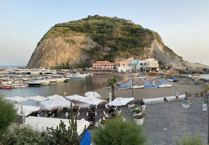 Il porto turistico di Ischia, con yacht ormeggiati, una spiaggia con lettini e ombrelloni, e case colorate ai piedi di una collina rocciosa.