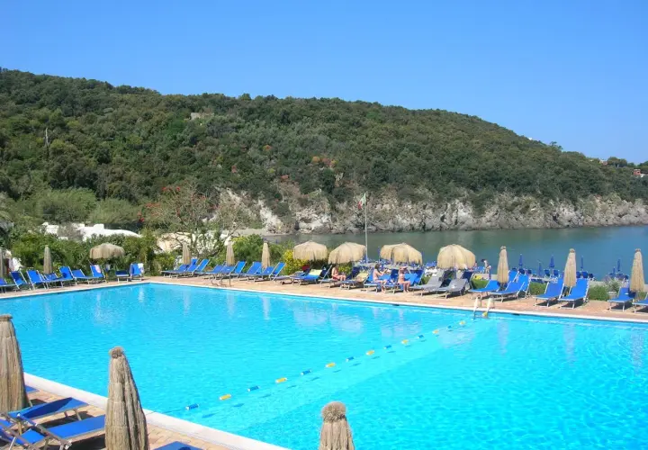 Piscina azzurra con lettini e ombrelloni, vista sul mare e collina verdeggiante. Un angolo di paradiso, forse Negombo o un luogo ispirato a Poseidon.