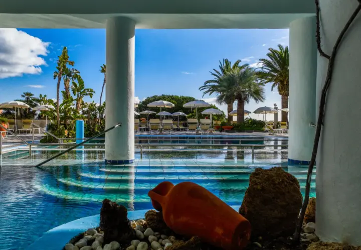 Piscina azzurra con palme e lettini, un'atmosfera da **parco termale romantica** con un'antica anfora come decorazione.