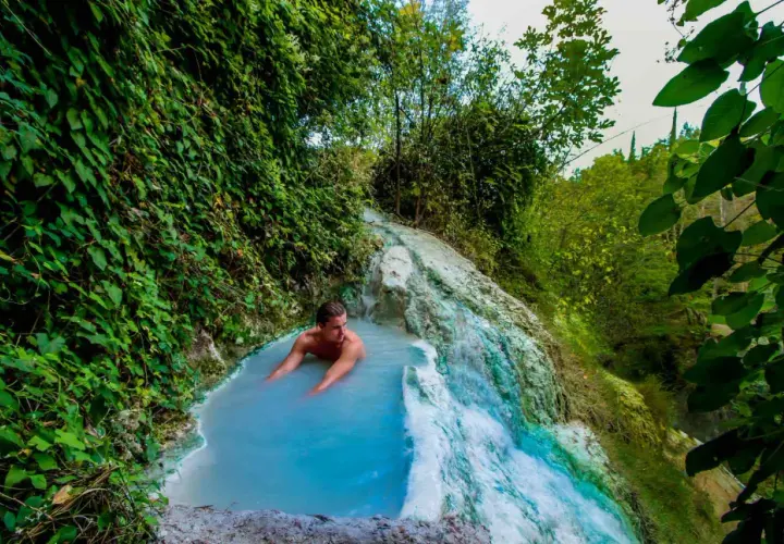 Un uomo si rilassa nelle acque azzurre di una cascata termale naturale a Olmitello, Ischia, circondato da lussureggiante vegetazione.