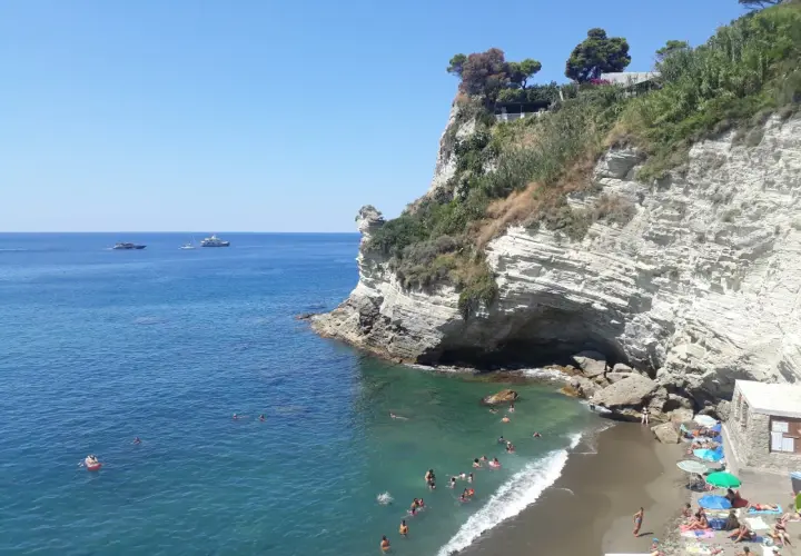 Scogliera bianca a picco sul mare turchese, con bagnanti e yacht all'orizzonte. La **cava grado Ischia** offre un rifugio naturale.