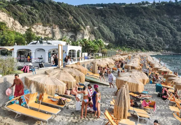 Spiaggia di Citara affollata, con ombrelloni di paglia, lettini arancioni e gente che si gode il mare. Per arrivare, segui le indicazioni per Forio.