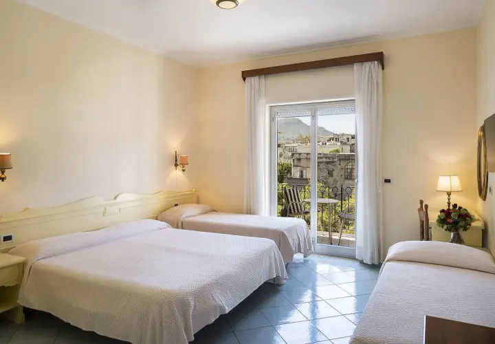 Camera con tre letti singoli, vista su un borgo di Alexander Ischia. Balcone con tavolo e sedie.
