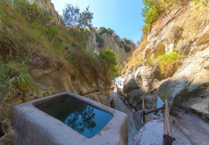 Piscina naturale scavata nella roccia, parte delle storiche terme di Cavascura, circondata da vegetazione e pareti rocciose.