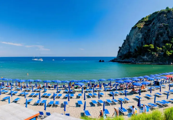 Spiaggia affollata con ombrelloni blu e lettini sulla riviera di San Montano. Mare azzurro e scogliera verde sullo sfondo.