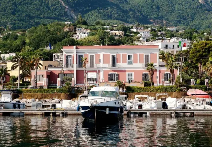 Il **hotel Svizzera Ischia** si affaccia sul porto con barche ormeggiate. Sullo sfondo, case arroccate su una collina verdeggiante.