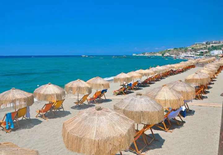 Spiaggia attrezzata con ombrelloni e lettini, mare azzurro e case in collina. Perfetto per chi cerca il **hotel San Valentino Ischia distanza dal mare** ideale.