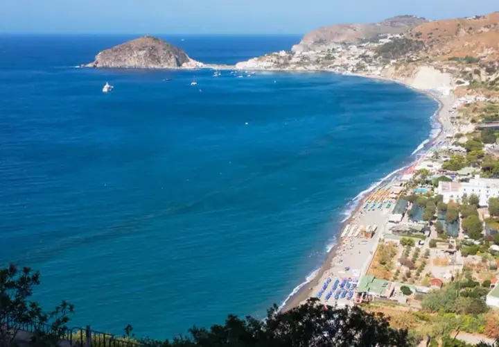 Vista panoramica della spiaggia dei Maronti con lidi attrezzati, ombrelloni colorati e il mare blu intenso. In lontananza, un isolotto e la costa frastagliata.