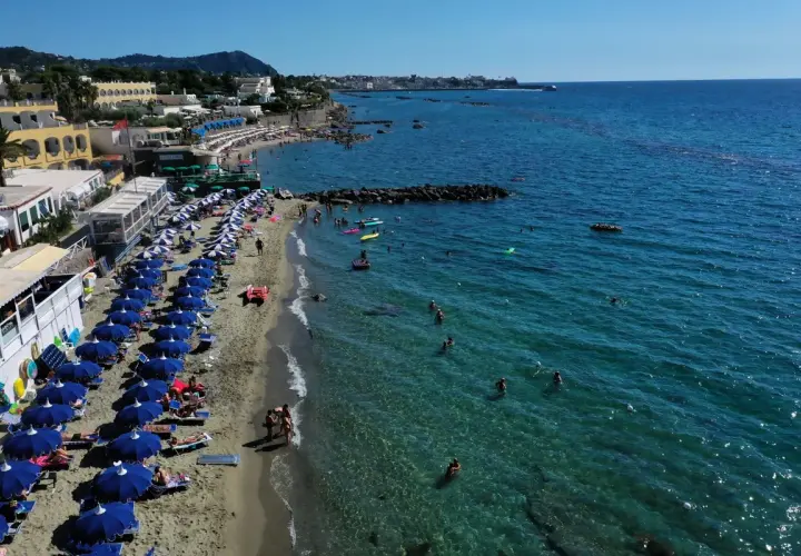 Forio Ischia spiagge affollate sotto il sole. Ombrelloni blu, bagnanti in mare e hotel affacciati sulla costa.