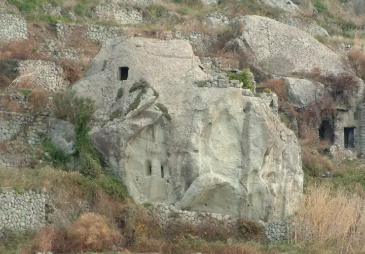 Antiche case in pietra scavate nella roccia e costruite con muretti a secco, immerse in un paesaggio collinare brullo.