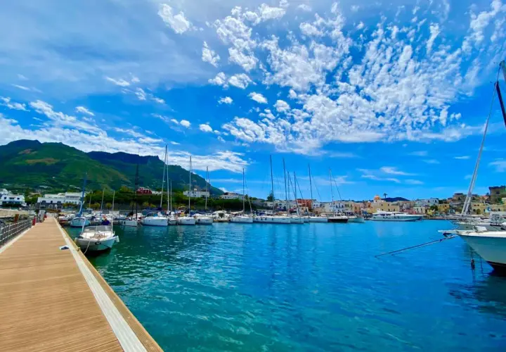Porto di Forio, Ischia: barche a vela ormeggiate in un mare azzurro sotto un cielo con nuvole bianche. Per raggiungere Forio da Ischia Porto, si può prendere un autobus o un taxi.