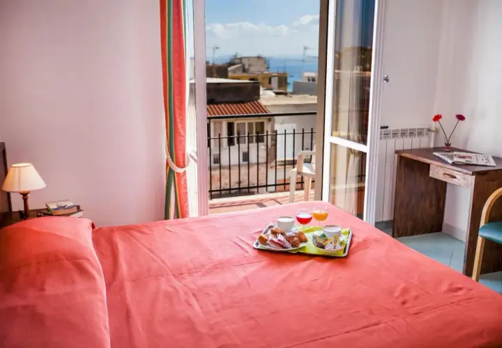 Camera con letto rosso e vassoio colazione sul letto. Vista mare dal balcone dell'hotel Conte Ischia.