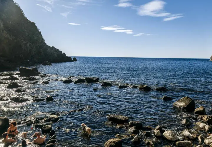Persone e un cane si godono le acque calde della **baia di Sorgeto**, tra scogli e rocce a picco sul mare blu.