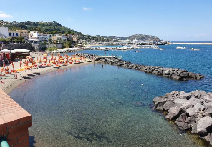 La spiaggia della marina è baciata dal sole, con ombrelloni arancioni e lettini pronti per accogliere i bagnanti. Il mare cristallino invita a un tuffo.