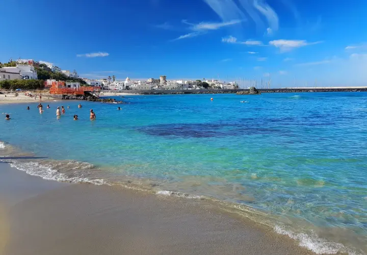 Persone nuotano nelle acque cristalline della spiaggia della Chiaia, con un borgo bianco sullo sfondo e un cielo azzurro con nuvole bianche.