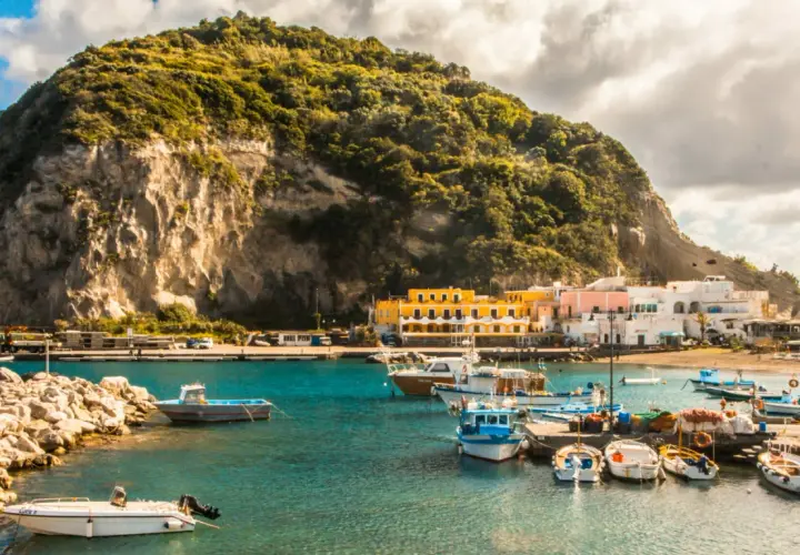 Un pittoresco ormeggio a Ischia, con barche colorate ormeggiate davanti a case bianche e gialle, sotto una collina verdeggiante.