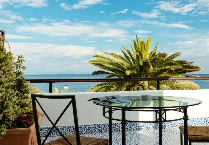 Vista mare da un beach club a Ischia: tavolino in vetro, sedia in ferro e palma rigogliosa sotto un cielo azzurro.