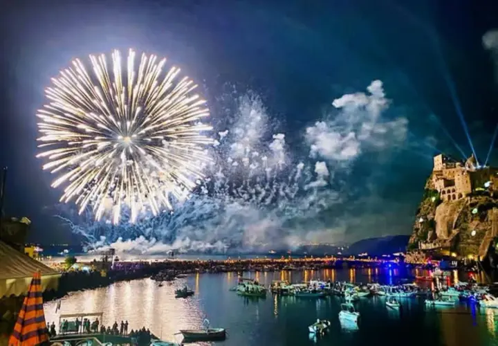 Fuochi d'artificio spettacolari illuminano il cielo notturno sopra un porto pieno di barche, con un castello arroccato su una scogliera. Un'immagine che ricorda le celebrazioni di Mario Mazzella.
