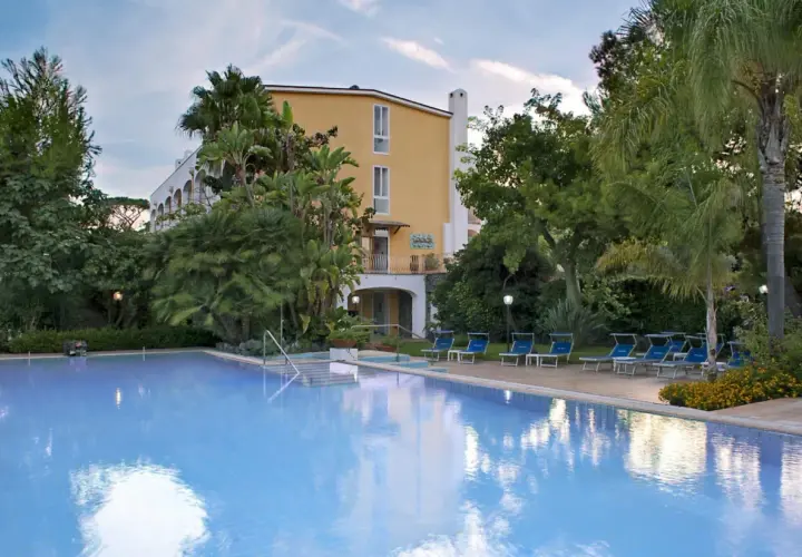 Piscina azzurra e lettini blu in un rigoglioso giardino tropicale, con un edificio giallo sullo sfondo, a San Giovanni, Ischia.