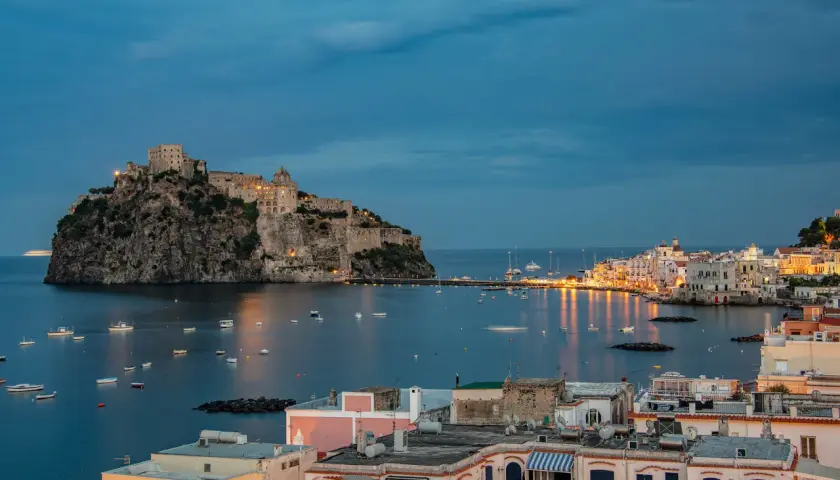Castello Aragonese domina il mare blu, con barche che ondeggiano e luci che illuminano le pittoresche calette di Ischia al crepuscolo.