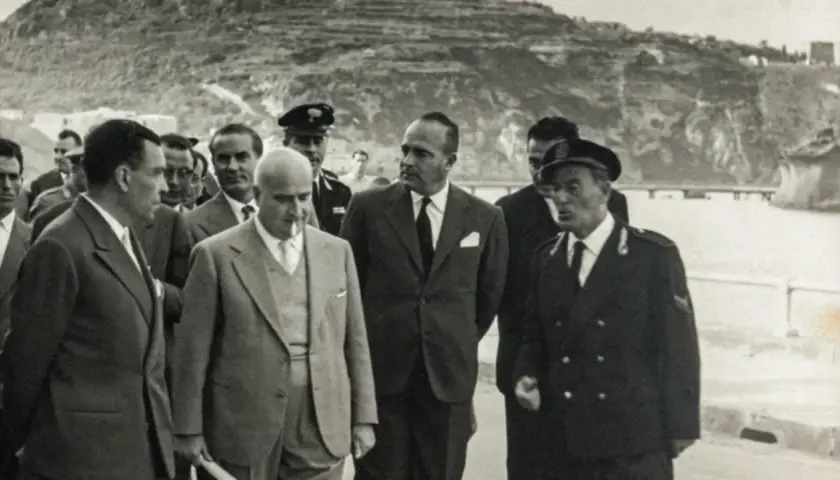 Angelo Rizzoli, con abito scuro, cammina con altri uomini a Ischia, sullo sfondo una collina terrazzata.