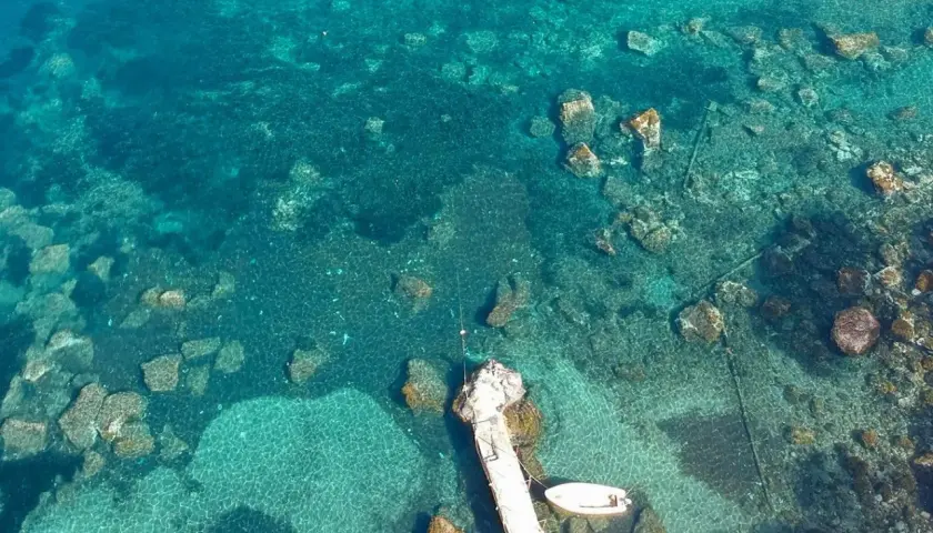 Acque cristalline e fondali rocciosi ad Aenaria, Ischia. Un pontile accoglie piccole imbarcazioni.