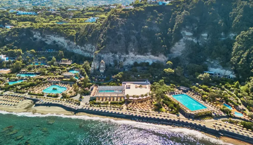 Vista aerea di un resort con piscine e lettini sulla spiaggia, incastonato tra il mare e una scogliera verdeggiante. Un paradiso di terme a Forio.