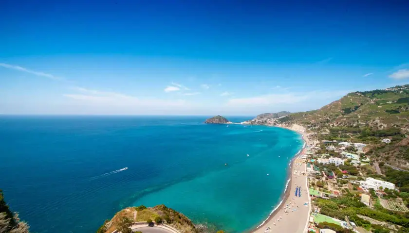 Vista panoramica delle spiagge libere a Ischia, con il mare turchese che lambisce la costa e le case arroccate sulle colline verdi.