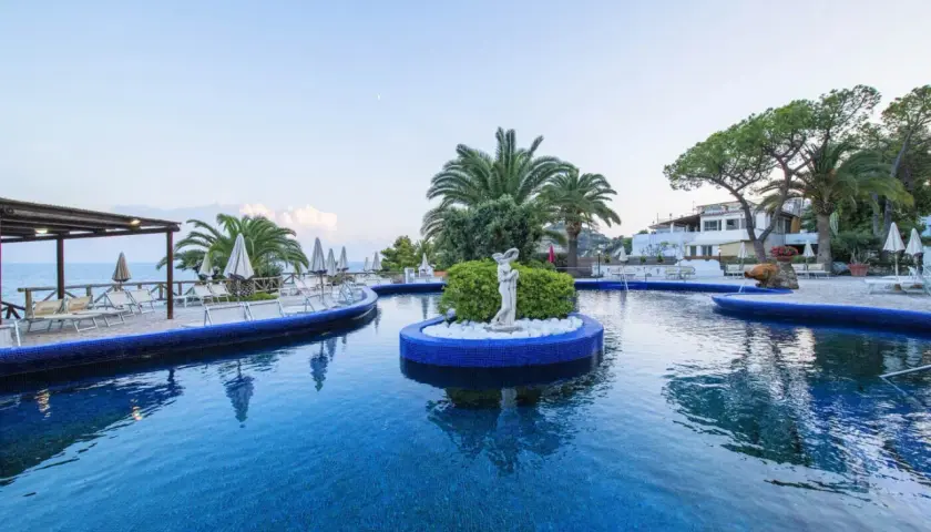 Piscina azzurra con isolotto centrale e statua, lettini e palme intorno. Un'oasi di relax all'Hotel Apollon.