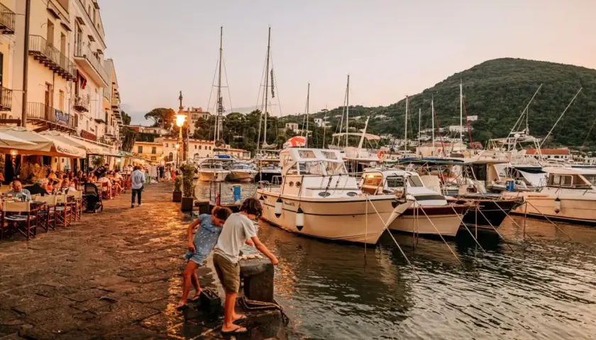 Bambini giocano sul molo della riva destra Ischia, con ristoranti affollati e barche ormeggiate al tramonto.