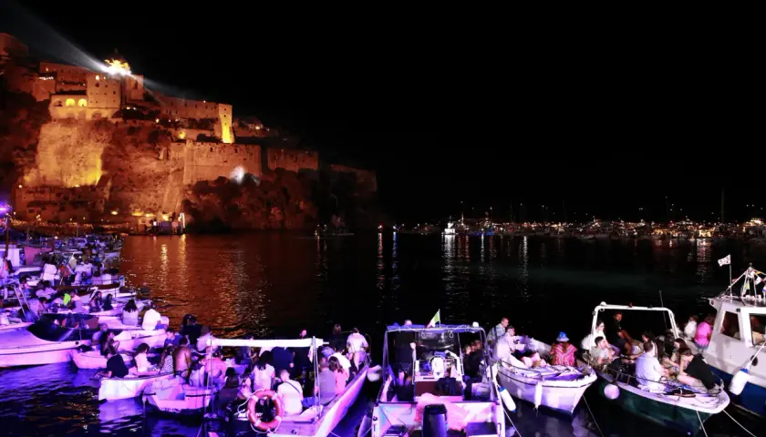 Una magica festa a Ischia: barche illuminate e gente festante con il castello illuminato sullo sfondo.