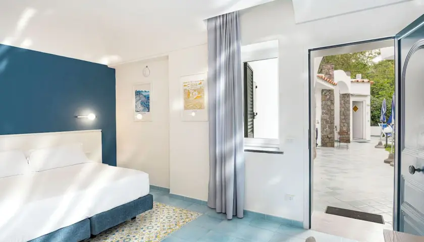 Camera luminosa con letto matrimoniale e vista su un cortile con ombrelloni blu. Un invito a soggiornare all'hotel oasi Ischia.