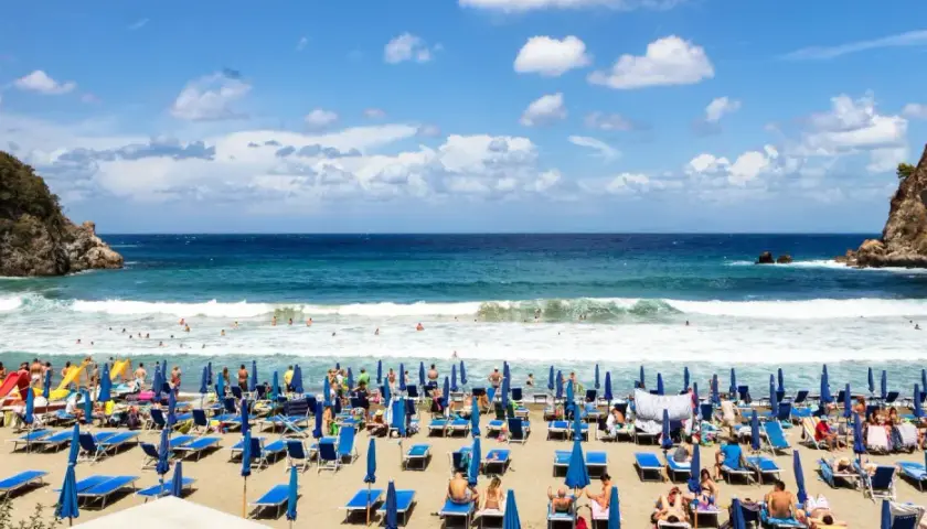 Ischia ad agosto: una spiaggia affollata con lettini blu, ombrelloni e bagnanti che si godono il mare cristallino sotto un cielo azzurro con nuvole bianche.