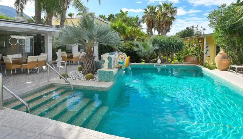 Piscina cristallina con cascata decorativa, lettini prendisole e area ristoro esterna. Un angolo di relax che ricorda le terme comunali di Ischia.