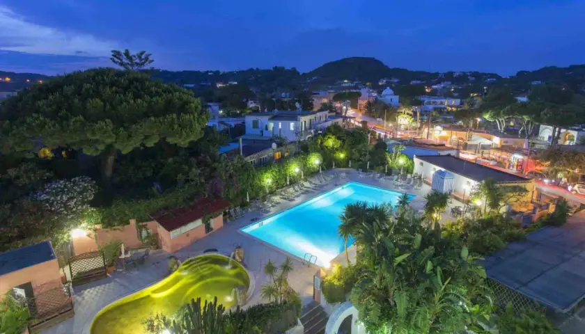 Piscina illuminata e vasca idromassaggio a forma di fagiolo al Hotel Eden Ischia, con vista sul paese di sera.
