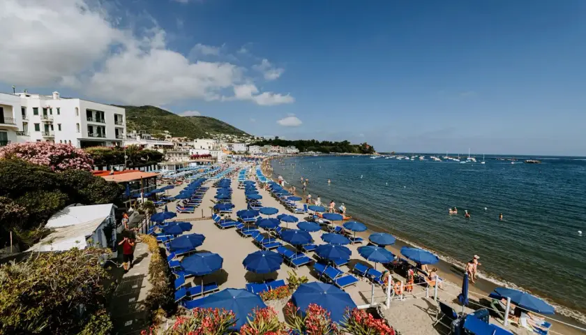 Spiaggia affollata a Ischia Lido con file di ombrelloni blu e lettini, bagnanti in acqua e barche all'orizzonte.