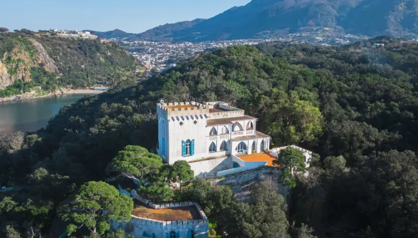 Villa La Colombaia a Ischia, un bianco maniero con torrette e finestre blu, immerso nel verde lussureggiante con vista sul mare e sulle montagne.
