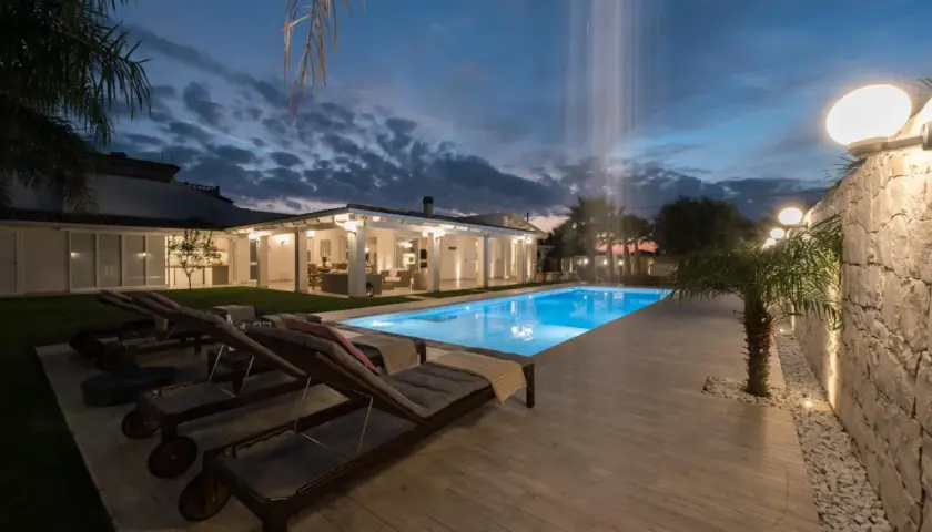 Piscina illuminata e lettini prendisole al tramonto, con cascata d'acqua. Un'oasi di relax al **hotel le palme Ischia**.