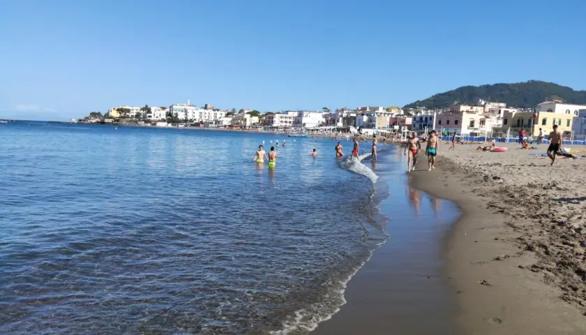 Persone si godono una giornata di sole sulla spiaggia di San Pietro, con il mare azzurro e case colorate sullo sfondo.