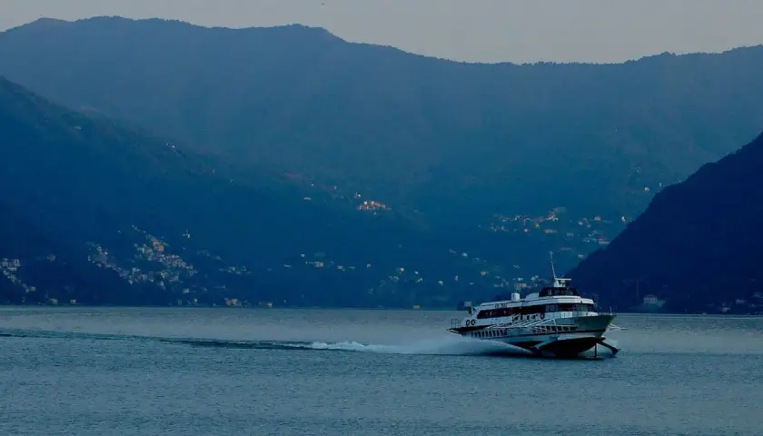 Aliscafo in navigazione su un lago calmo, con montagne boscose e case illuminate sullo sfondo.