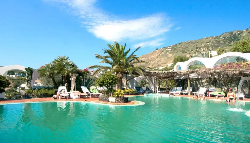 Piscina con lettini e palme, un'oasi di relax alle terme Sant'Angelo Ischia, con vista sulla collina verdeggiante.