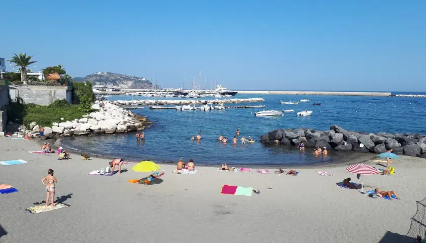Spiaggia della Marina di Ischia: bagnanti si godono il sole e il mare, con barche ormeggiate nel porto sullo sfondo.