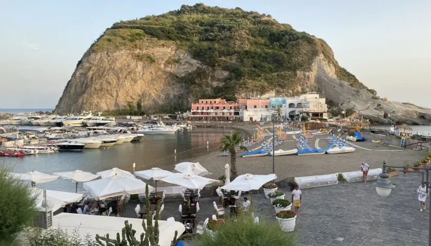 Il porto turistico di Ischia, con yacht ormeggiati, una spiaggia con lettini e ombrelloni, e case colorate ai piedi di una collina rocciosa.