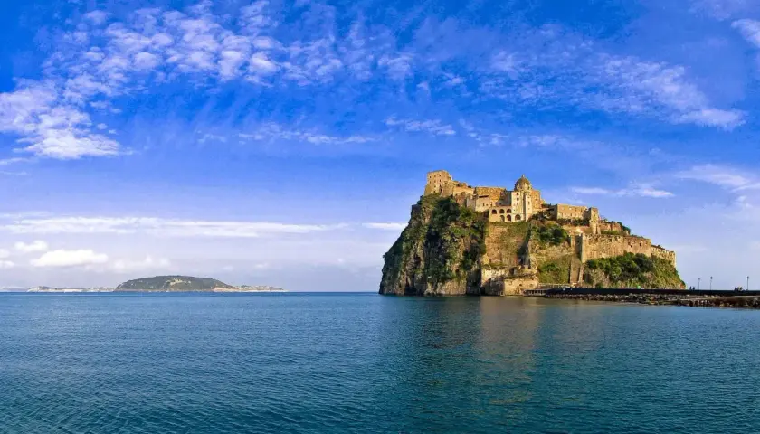 Il **palazzo reale** di Ischia, arroccato su uno scoglio, domina il mare blu sotto un cielo terso.