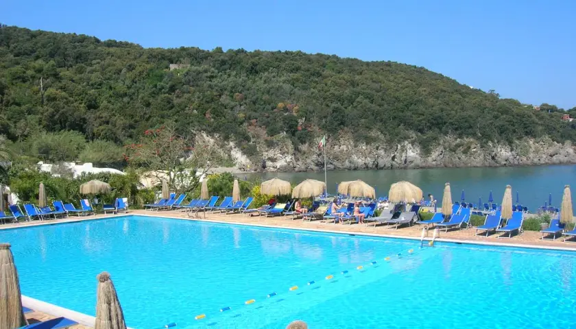 Piscina azzurra con lettini e ombrelloni, vista sul mare e collina verdeggiante. Un angolo di paradiso, forse Negombo o un luogo ispirato a Poseidon.