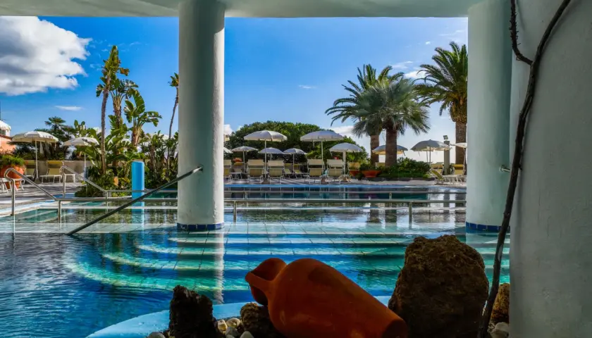 Piscina azzurra con palme e lettini, un'atmosfera da **parco termale romantica** con un'antica anfora come decorazione.